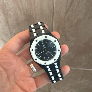 May include: A black and white wristwatch featuring an octagonal bezel with eight visible screws. The black dial displays the text AP, Swatch, and Royal Rip, with white hour markers and hands. The integrated strap is black with white centre links.