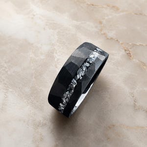 Può includere: Un anello nero con superficie esterna sfaccettata e opaca, caratterizzato da un intarsio centrale in materiale argentato brillante frantumato. L'interno della fascia è liscio e lucido, con una finitura argento metallizzato che contrasta con la texture scura.