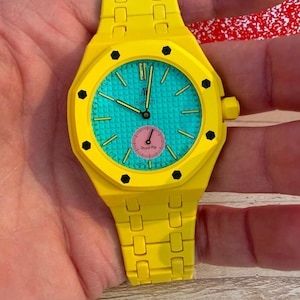 Può includere: Un orologio da polso giallo brillante con lunetta ottagonale e viti esagonali nere. Il quadrante è turchese testurizzato con indici gialli, lancette nere e un piccolo sotto-quadrante circolare rosa con la scritta Royal Pop e il logo Swatch.
