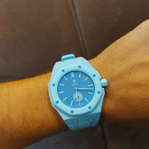 Puede incluir: Un reloj de pulsera azul claro con un bisel octogonal de ocho tornillos visibles, una esfera texturizada con las letras AP y la palabra Swatch, manecillas luminosas y una correa de silicona azul claro a juego con cierre de hebilla.