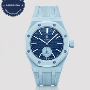 Könnte beinhalten: Eine hellblaue Armbanduhr mit dunkelblauem Zifferblatt und weißen Zeigern. Die Uhr hat ein strukturiertes Armband und ein rundes Zifferblatt mit dem Text "AP swatch" und "Royal Pop". Das Bild enthält auch eine kreisförmige Grafik mit "Straps for Royal Pop" und "COMING SOON".
