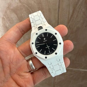 Puede incluir: Un reloj de pulsera blanco con una esfera circular negra que presenta un patrón de rejilla texturizado. La esfera muestra el texto AP Swatch y Royal Oak. Tiene una correa de eslabones blanca y un bisel octogonal blanco asegurado con ocho tornillos hexagonales negros.