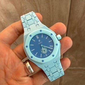 Puede incluir: Un reloj de pulsera azul claro con un bisel octogonal y ocho tornillos visibles. La esfera es de color azul oscuro con un patrón de rejilla texturizado, mostrando los logotipos AP y Swatch, manecillas plateadas y una pequeña subesfera circular.