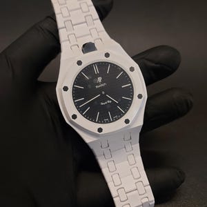 Puede incluir: Un reloj de pulsera blanco con una esfera negra que presenta un patrón de cuadrícula texturizado. La esfera muestra el texto AP, Swatch y Royal Rop, con marcadores de hora y manecillas negras. El bisel octogonal tiene ocho tornillos negros y la correa es blanca.