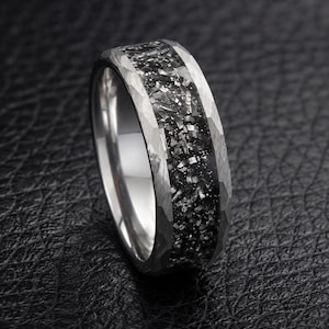 Può includere: Un anello color argento con finitura in metallo martellato sui bordi e un ampio inserto centrale incassato, riempito con un materiale scuro, testurizzato e scintillante che ricorda frammenti di pietra o metallo in nero e grigio.