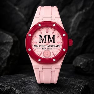 Op de afbeelding: Een lichtroze polshorloge met een donkerrode achthoekige ring voorzien van zilveren schroeven. De roze wijzerplaat toont de tekst MM, MM CUSTOM STRAPS en NEW YORK, met een kleine sub-wijzerplaat onderaan en een bijpassende roze rubberen band.