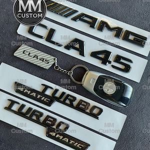 May include: Black AMG, CLA 45, and Turbo 4MATIC emblems, along with a car key and a CLA 45 keychain. The emblems are glossy and mounted on white strips. The key has a silver and black design.