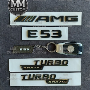 Puede incluir: Emblemas de coche negros AMG, E53, Turbo y 4MATIC y un llavero. Los emblemas están sobre un respaldo blanco. El llavero tiene un acabado plateado y un logotipo de Mercedes-Benz. La imagen también incluye el texto "MM Custom".