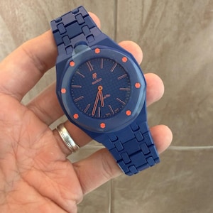 May include: A blue wristwatch featuring a textured dial with orange hour markers and hands. The watch has a blue integrated link bracelet and a blue bezel with orange accents. The dial displays the text AP, Swatch, and Royal Oak in orange.
