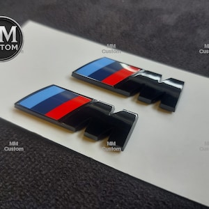 Pode incluir: Dois emblemas "M" pretos e cromados com listras azuis, vermelhas e brancas. Os emblemas estão em uma superfície branca com o logotipo "MM Custom" no canto superior esquerdo e repetido no fundo.