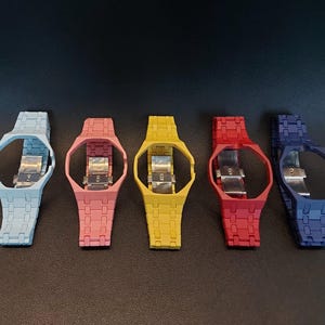 Op de afbeelding: Vijf horlogebanden met geïntegreerde behuizingen op een rij, met een matte afwerking in lichtblauw, roze, geel, rood en donkerblauw. Elke band bevat een zilverkleurig metalen vouwsluitingsmechanisme dat in het midden zichtbaar is.