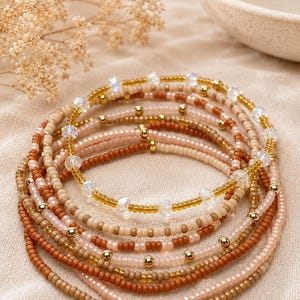 Puede incluir: Un conjunto de pulseras apiladas con pequeñas cuentas redondas en tonos beige, bronceado y marrón, intercaladas con esferas de oro metálico y cuentas de cristal transparente facetadas dispuestas en un patrón circular.