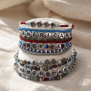 Puede incluir: Una pila de pulseras de cuentas con las palabras "LOST" y "AMERICANA" deletreadas con cuentas de letras plateadas. Las pulseras presentan cuentas rojas, blancas, azules y plateadas. Las pulseras están apiladas sobre una superficie blanca.