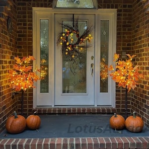 May include: A white front door with a glass panel and an arched transom window, decorated with a twig wreath featuring orange lights. Two illuminated artificial maple trees with orange leaves flank the door, with two pumpkins on each side.