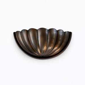 May include: A bronze-colored, half-circle wall sconce with a fluted, shell-like design. The sconce has a dark brown trim and is mounted on a white wall. The fluted design creates a series of vertical, curved ridges.