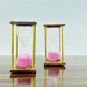 May include: Two hourglasses with pink sand, clear glass, and wooden bases. The hourglasses have a dark wood top and bottom, with gold-colored metal supports. The sand is a vibrant pink color, and the glass is clear.