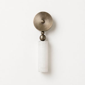May include: A wall sconce featuring a circular, antique brass fixture above a white, cylindrical base. The brass element includes a disc and a sphere, adding a touch of elegance. The sconce is designed for interior lighting and adds a decorative element.