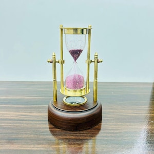 May include: An hourglass with a brass frame and a wooden base. The glass contains pink sand. The hourglass is a decorative item, likely used for timing or as an ornamental piece. The base is a dark brown color.