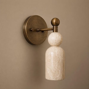 May include: A wall-mounted light fixture featuring a circular antique brass backplate and a horizontal arm. The lamp includes a spherical brass finial, a round stone orb, and a cylindrical stone shade, all showcasing a natural beige marble pattern.