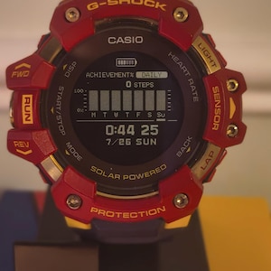 May include: A G-Shock Casio solar-powered digital watch with a red and yellow bezel. The circular display shows a battery icon, achievement progress, 0 steps, a weekly bar graph, the time 0:44 25, and the date 7/26 SUN. Buttons are labeled.