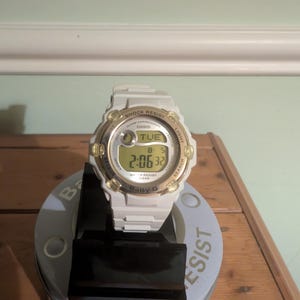 Puede incluir: Reloj digital Baby-G blanco con detalles dorados. El reloj muestra el día, la fecha y la hora. Es resistente a los golpes y al agua. El reloj está sobre un soporte negro.