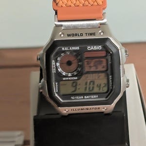 May include: A silver Casio digital watch with an orange woven strap. The watch face displays the time, world time, and other functions. The text "WORLD TIME", "CASIO", and "ILLUMINATOR" are visible on the watch.