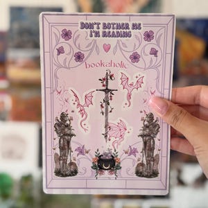 May include: A rectangular sticker sheet with a light purple background featuring the text DON'T BOTHER ME I'M READING and bookaholic. The design includes two suits of armor, a central sword entwined with vines, three small pink dragons, and a cauldron.