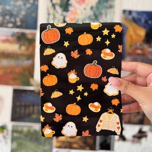 Pode incluir: Uma bolsa retangular preta com um padrão repetitivo de abóboras cor de laranja, fantasmas brancos, estrelas amarelas, folhas de outono, fatias de tarte de abóbora, canecas com natas batidas e um casaco de malha creme com corações vermelhos.