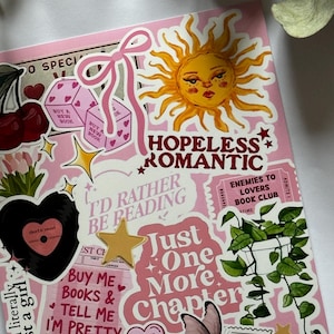 Op de afbeelding: Een roze oppervlak bedekt met diverse stickers, waaronder een gele zon, een roze strik, kersen, dobbelstenen, een zwarte vinylplaat, een kamerplant, een vlinder en tekst met "HOPELESS ROMANTIC", "I'D RATHER BE READING" en "Just One More Chapter".