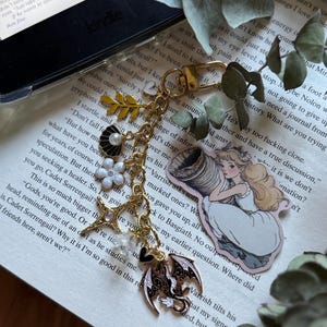 Pode incluir: Um porta-chaves de metal dourado com vários berloques, incluindo uma folha amarela, uma concha preta e branca com uma pérola, uma flor branca, uma estrela, um pequeno coração e um dragão preto e branco, com uma ilustração de um elfo loiro.