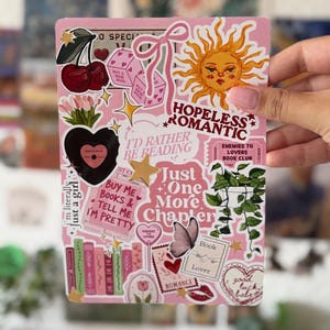 Op de afbeelding: Een rechthoekig roze stickervel met diverse illustraties waaronder een zon, kersen, roze dobbelstenen, een zwart hart, een kamerplant, een vlinder en boeken. Tekst bevat onder andere "Hopeless Romantic" en "Just One More Chapter".