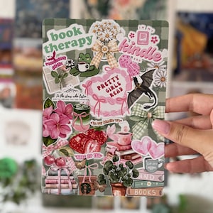 May include: A rectangular sticker sheet featuring a sage green checkered background covered in various illustrations including pink flowers, stacks of books, a green frog with a coffee cup, a black dragon, strawberries, and text reading book therapy.