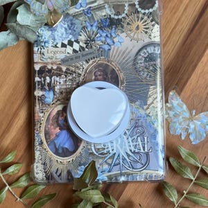 Può includere: Una custodia trasparente per telefono con un design a collage con illustrazioni vintage, farfalle e la parola "CARAVAL". È attaccato un supporto per telefono bianco a forma di cuore. Foglie decorative e una superficie in legno completano la scena.