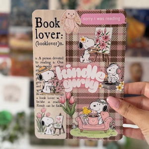 Peut inclure: Un autocollant rectangulaire présentant une définition de dictionnaire pour "book lover", des illustrations de Snoopy, et le texte "kindle girly" et "sorry i was reading". Le design inclut un motif écossais marron, des tulipes roses et un lapin.