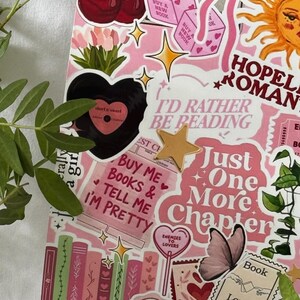 Op de afbeelding: Een roze collage met diverse stickers waaronder kersen, dobbelstenen, een zon, een zwart hart en boeken. De tekst bevat "I'd Rather Be Reading", "Just One More Chapter", "Buy Me Books & Tell Me I'm Pretty", "Enemies to Lovers" en "Book Lover".