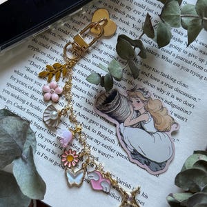 May include: A gold-toned metal keychain featuring a heart-shaped clasp and a series of charms including a gold leaf, a pink flower, a white shell with a pearl, a pink bell-shaped flower, a pink daisy, a white butterfly, a pink heart with wings, and a star, alongside a sticker of a blonde character in a white dress holding a large horn.