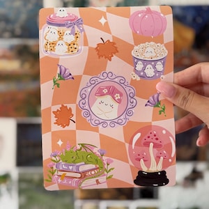 Pode incluir: Uma folha de autocolantes retangular com um fundo axadrezado em pêssego e branco, apresentando várias ilustrações, incluindo um frasco de bolachas de fantasmas, uma abóbora, um balde de pipocas com fantasmas, um fantasma emoldurado, folhas de outono, um dragão sobre livros e cogumelos.
