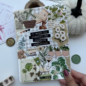 Puede incluir: Un diario decorado con varias pegatinas, incluyendo diseños florales y botánicos, un ciervo y el texto "I work to support my reading addiction." Otras pegatinas incluyen temas relacionados con el café y los libros.