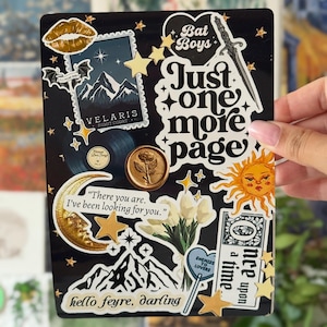 Könnte beinhalten: Eine schwarze rechteckige Tafel, die mit verschiedenen Aufklebern bedeckt ist, darunter ein goldener Lippenabdruck, eine Velaris-Briefmarke, ein Schwert, eine Sonne, ein Mond, ein Strauß weißer Tulpen und Text mit Bat Boys, Just one more page, There you are, hello feyre, darling.