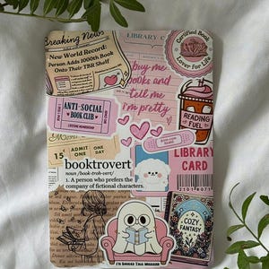 Könnte beinhalten: Ein rosa-beiges Buchcover mit Aufklebern und Text. Aufkleber sind Herzen, eine Kaffeetasse und die Wörter "booktrovert" und "Anti-Social Book Club". Das Cover hat ein "Cozy Fantasy"-Thema.