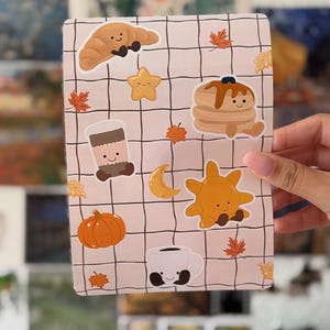 Puede incluir: Una hoja de pegatinas rectangular con un fondo de cuadrícula rosa claro que presenta varias ilustraciones de dibujos animados lindos, incluyendo un croissant, una estrella, tortitas, una taza de café, una luna creciente, un sol, una calabaza y pequeñas hojas de otoño naranjas.