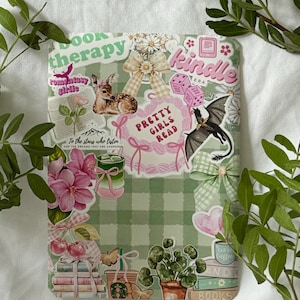 Könnte beinhalten: Eine grün-weiß karierte Kindle-Hülle, verziert mit Aufklebern. Aufkleber zeigen ein Reh, einen Drachen, Blumen, Schleifen, Würfel und die Texte "Pretty Girls Read" und "Book Therapy". Grüne Blätter umrahmen die Hülle.