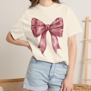 Può includere: Una t-shirt a maniche corte color crema con una grande illustrazione centrale in stile acquerello di un fiocco color rosa polvere. La maglietta presenta un girocollo e una vestibilità rilassata, abbinata a pantaloncini di jeans azzurri con risvolto.
