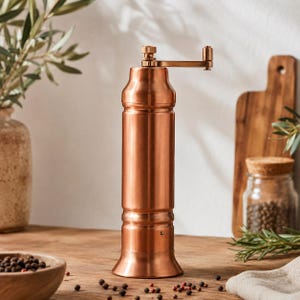 May include: A cylindrical copper-colored manual spice grinder featuring a metallic finish, a flared base, and a top-mounted crank handle. The body has a smooth, polished surface with subtle horizontal detailing near the top and bottom sections.