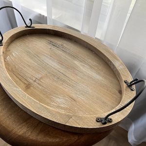 May include: Oval wooden serving tray with dark metal handles. The tray has a light wood finish with visible grain and a slightly distressed look. The handles are attached with small, dark metal brackets.