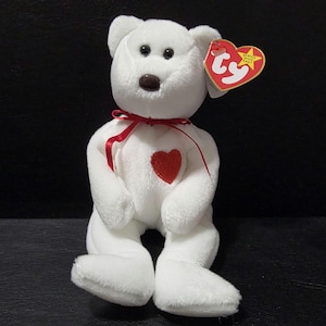 May include: A white plush teddy bear with a red heart on its chest and a red ribbon tied around its neck. The bear has black eyes and a brown nose. A red heart-shaped tag is attached to its ear.