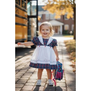 Peut inclure: Une jeune enfant porte une robe à carreaux avec un tablier blanc et un col blanc, tenant un sac à dos coloré. La robe a des accents rouges et bleus. L'enfant porte des baskets blanches et se tient sur une allée pavée.