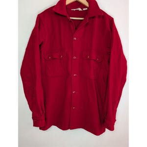 May include: A bright red button-down shirt with two chest pockets. The long-sleeved shirt is made of a thick material and has a classic collar. The shirt is hanging on a wooden hanger against a white background.