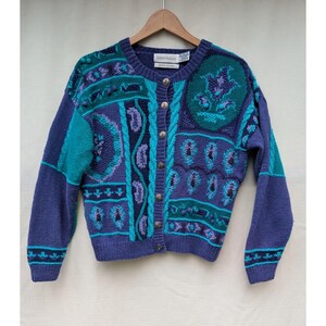 May include: A long-sleeve knit cardigan in shades of purple and teal, featuring a button-front closure with textured metal buttons, a round neckline, and intricate patterns including paisley motifs, cable knit details, and small pom-pom accents.