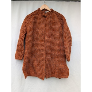 May include: A long, rust-coloured knitted coat with a high collar and three-quarter sleeves. The coat has a textured, woven appearance and fastens with two small, round buttons. The garment is hanging on a wooden hanger against a white backdrop.